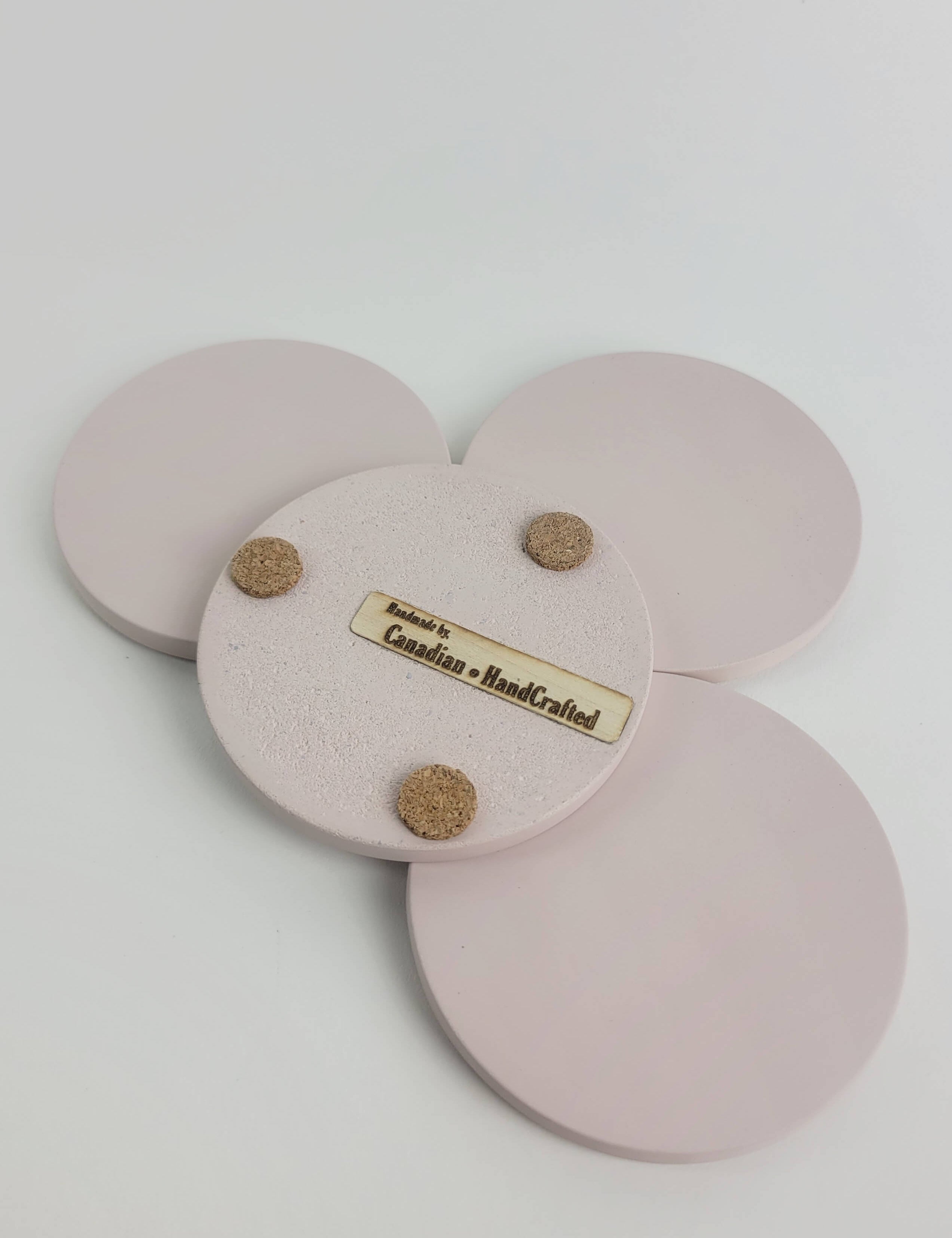 Set of 4 pastel pink concrete coasters, one flipped to reveal the underside featuring 3 cork bumpers and an engraved wooden veneer with the text 'Handmade by Canadian HandCrafted'.