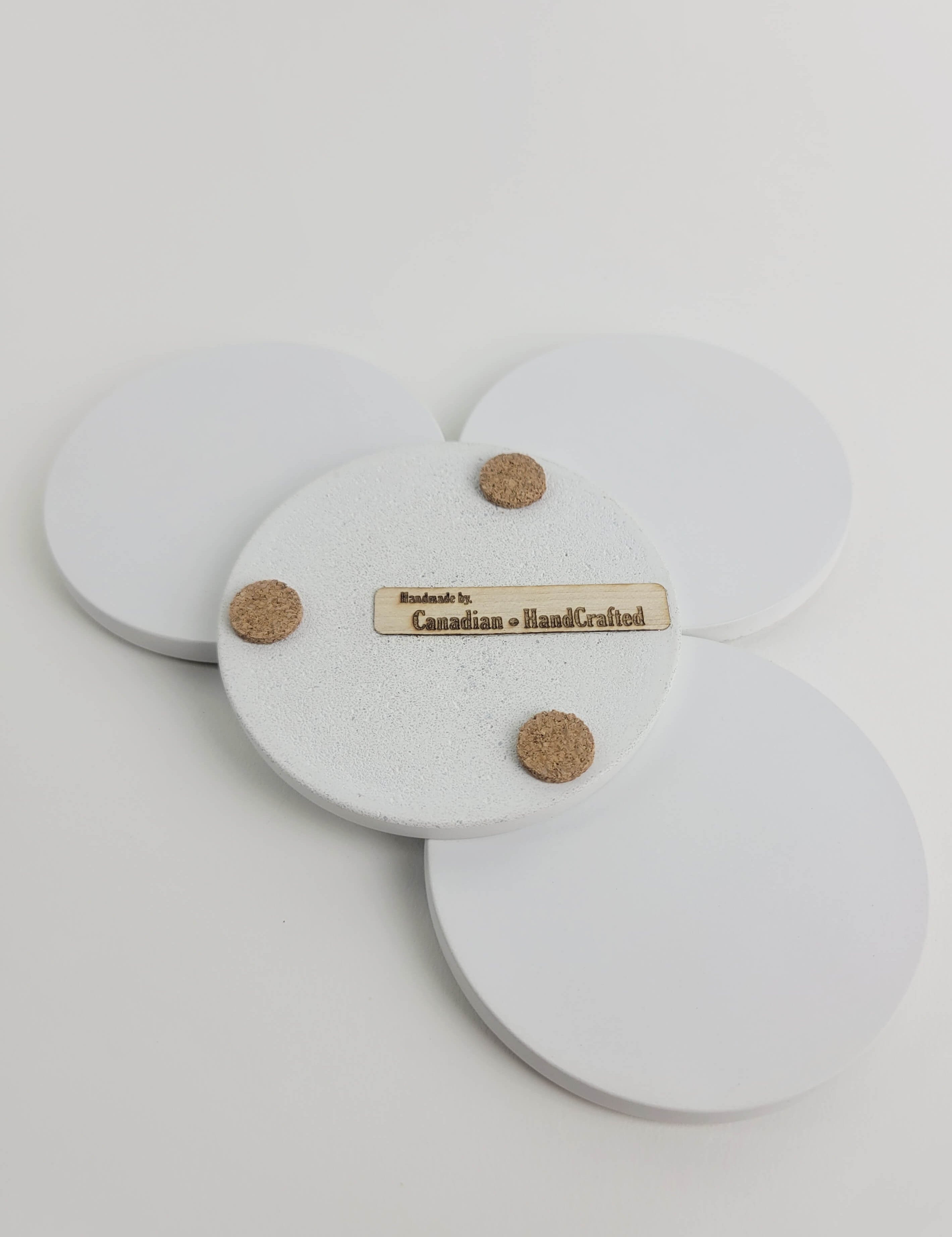 Set of 4 chalk white concrete coasters, one flipped to reveal the underside featuring 3 cork bumpers and an engraved wooden veneer with the text 'Handmade by Canadian HandCrafted'.