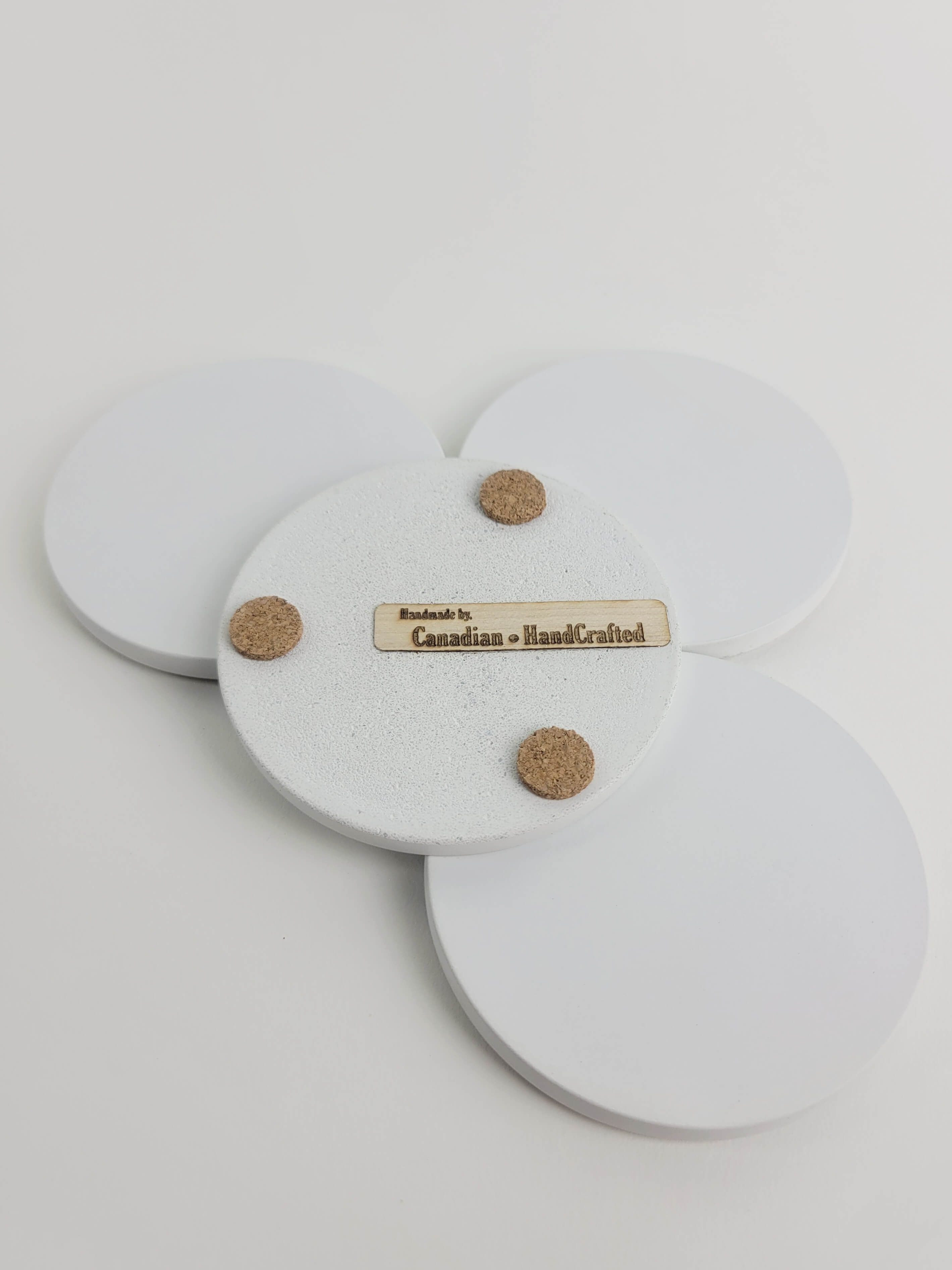 Set of 4 chalk white concrete coasters, one flipped to reveal the underside featuring 3 cork bumpers and an engraved wooden veneer with the text 'Handmade by Canadian HandCrafted'.