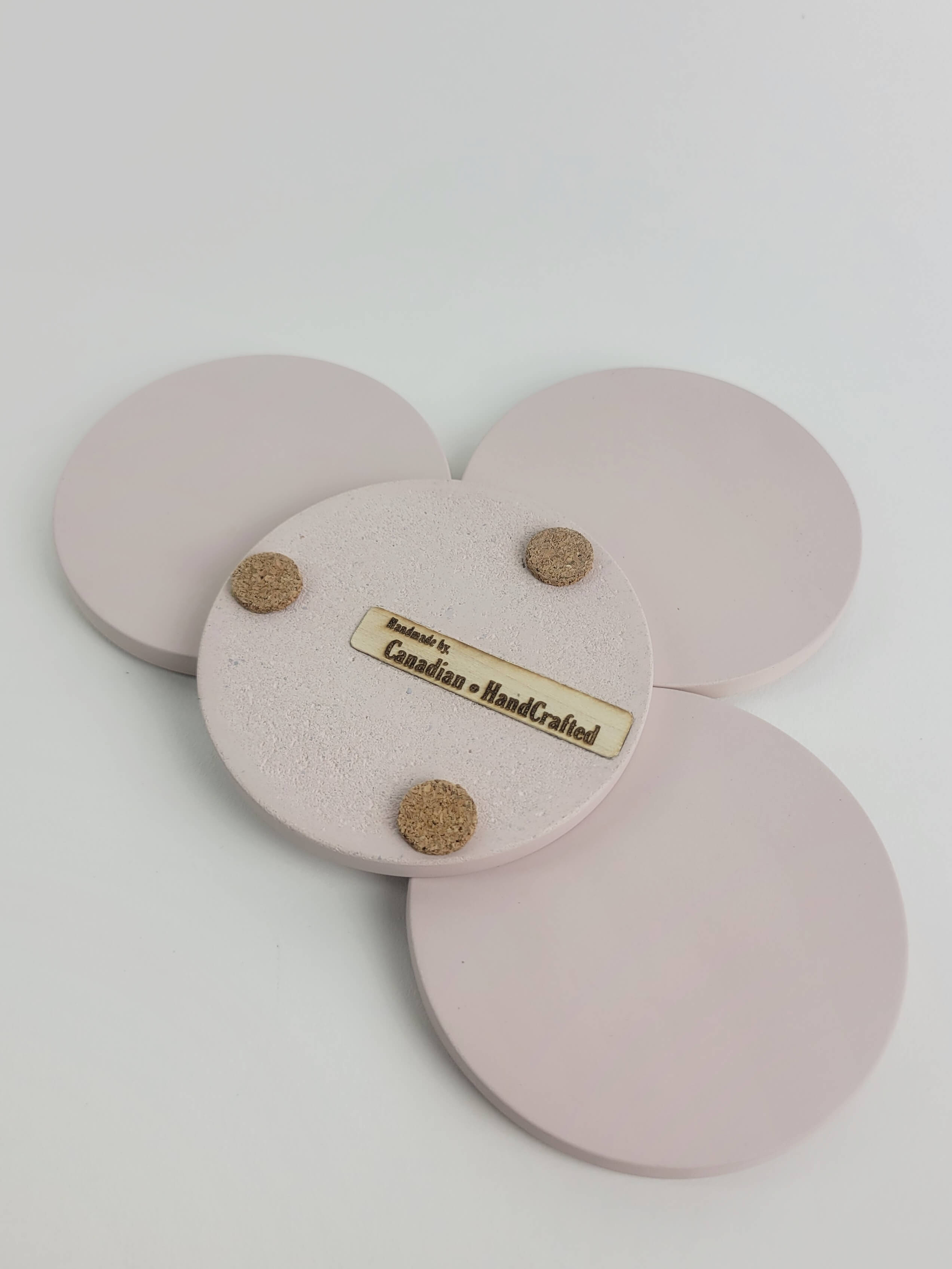 Set of 4 pastel pink concrete coasters, one flipped to reveal the underside featuring 3 cork bumpers and an engraved wooden veneer with the text 'Handmade by Canadian HandCrafted'.