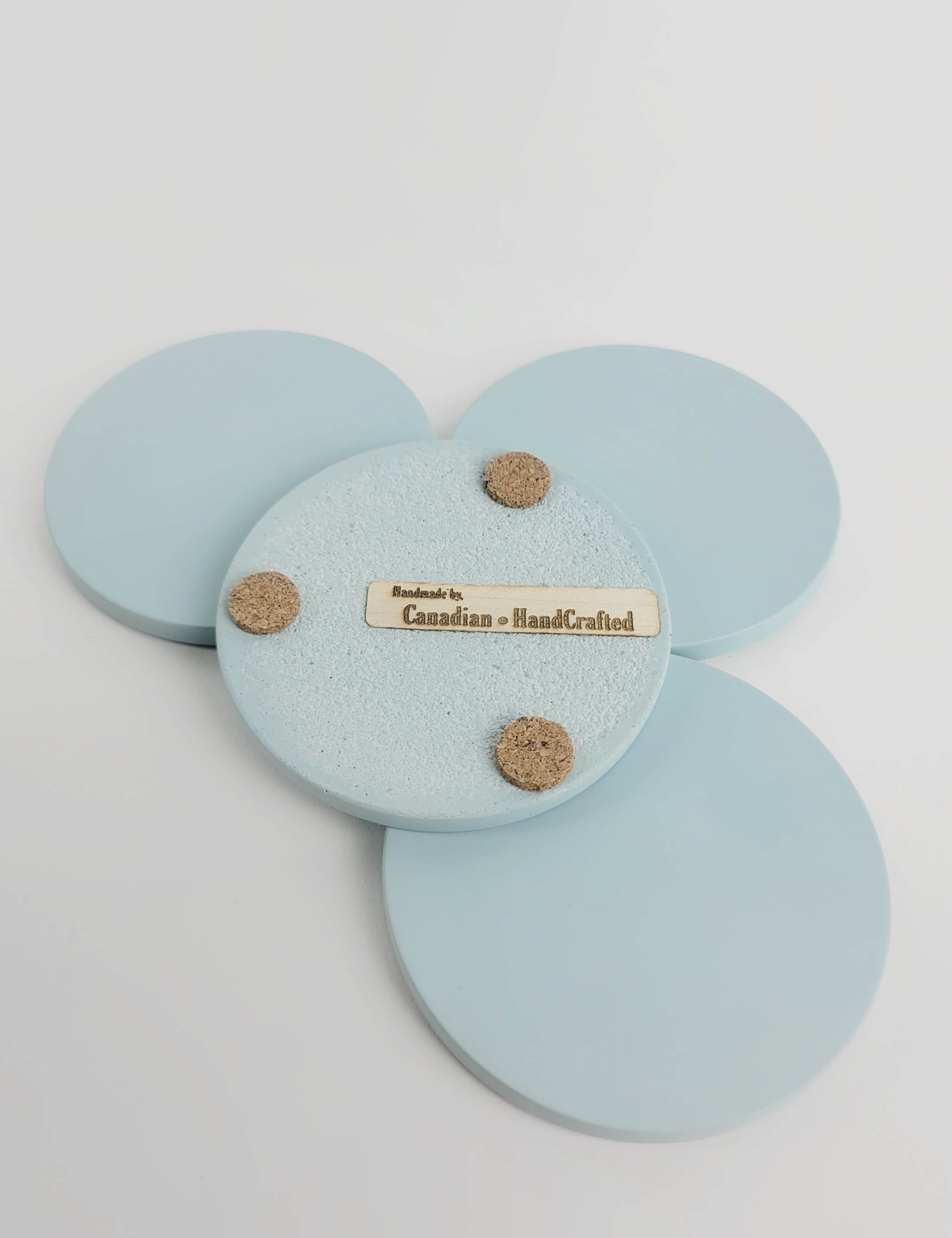 Set of 4 pastel turquoise concrete coasters, one flipped to reveal the underside featuring 3 cork bumpers and an engraved wooden veneer with the text 'Handmade by Canadian HandCrafted'.