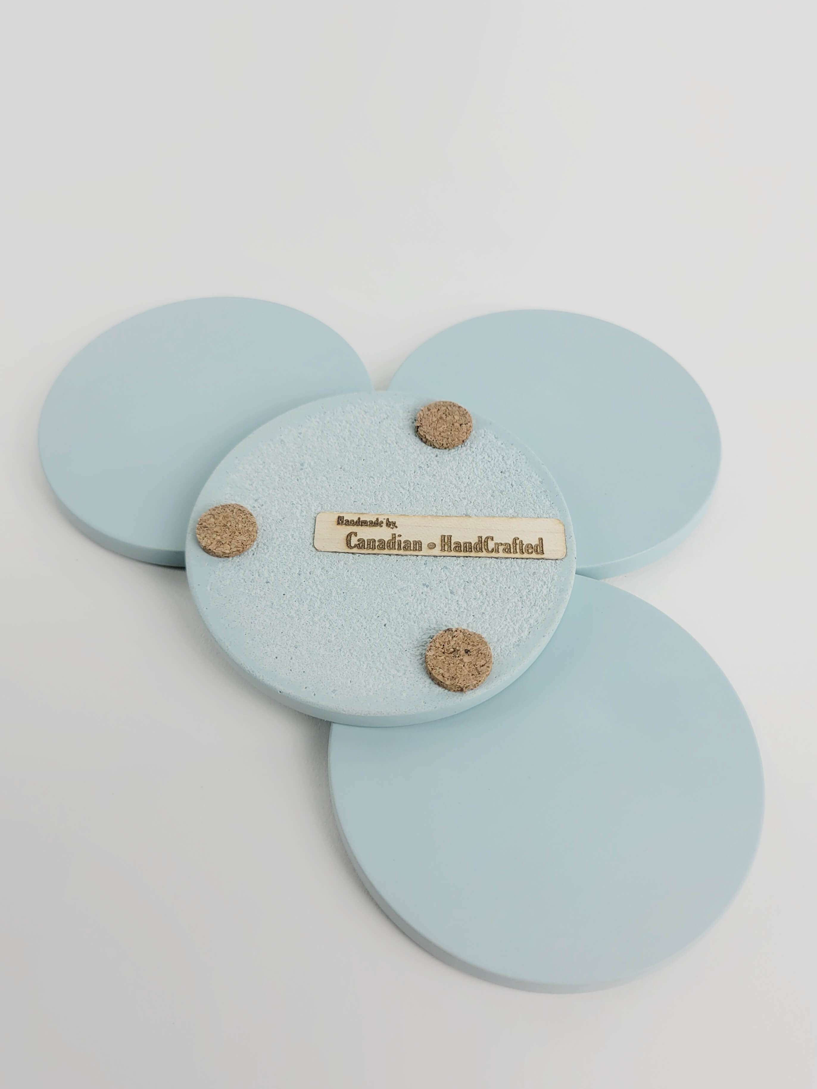 Set of 4 pastel turquoise concrete coasters, one flipped to reveal the underside featuring 3 cork bumpers and an engraved wooden veneer with the text 'Handmade by Canadian HandCrafted'.