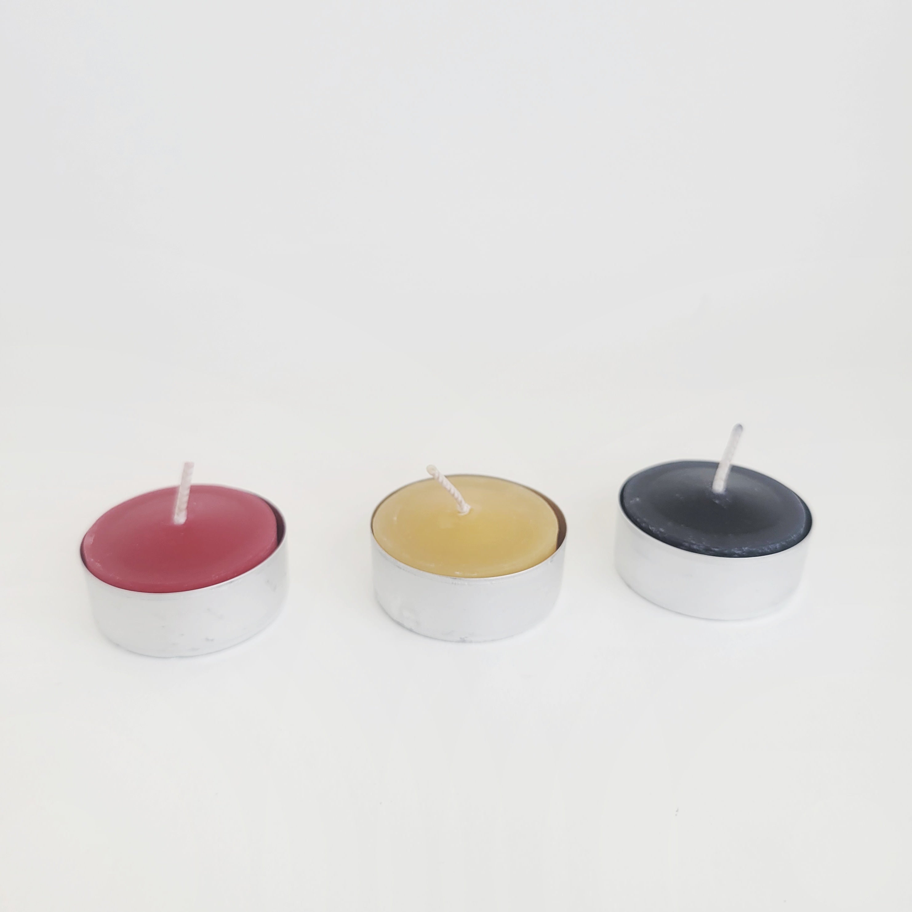 Colorful beeswax tealights - red, natural honey, and black - arranged side by side against a white background, perfect for home decor and ambiance.