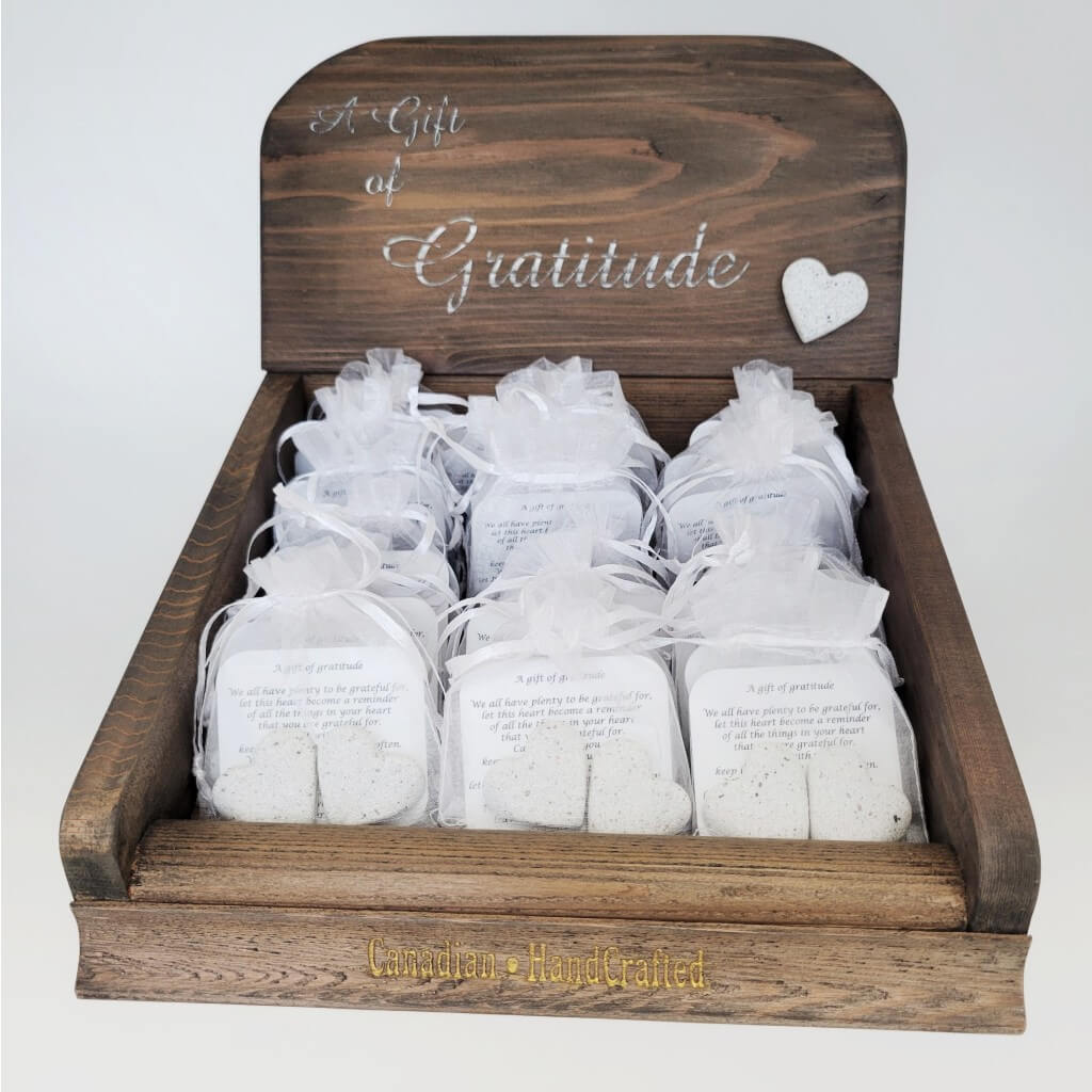 Enhance your display with a Gratitude Gift Pre-Pack Counter Display crafted from rich dark stained wood. Adorned with silver lettering engraved with 'a gift of gratitude', featuring a white stone heart affixed to the front. The display holds 24 silver translucent baggies, each containing two stone hearts and two gratitude notes. Spot a gold engraving at the base proclaiming 'Canadian HandCrafted'.