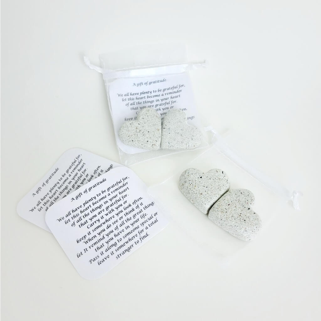 Two Gifts of gratitude, two stone hearts with two notes inside a translucent silver baggie. Another set of stone hearts and note is displayed below removed from the baggie.