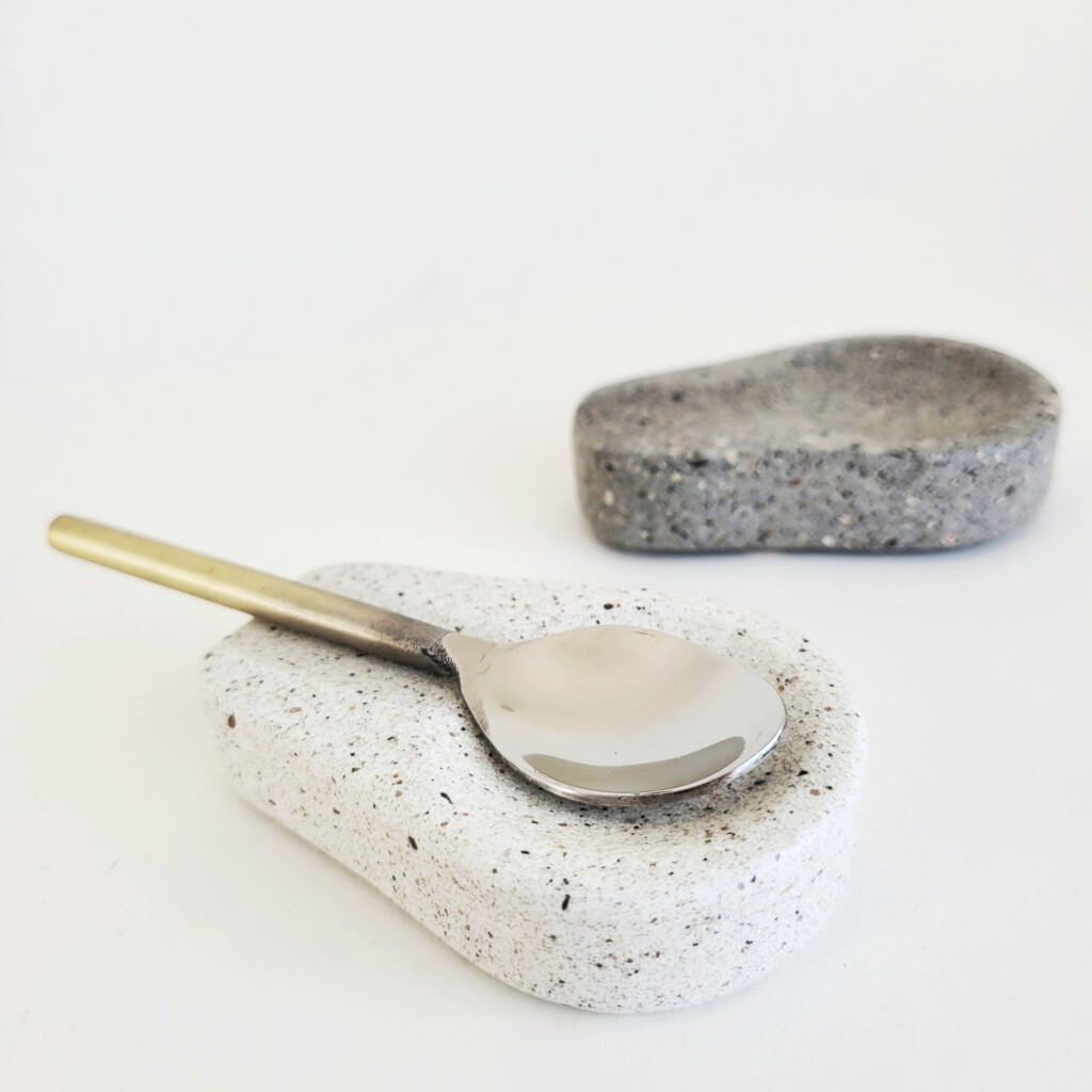 A white and grey terrazzo sandstone spoon rest. A gold and silver spoon rests on the white stone spoon holder.