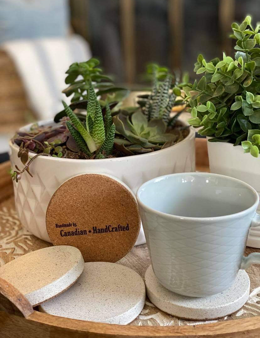 Set of 4 sparkling white sandstone coasters, one elegantly displayed on its edge to highlight the signature Canadian HandCrafted engraving on the cork bottom, complemented by beautiful green plants in the background.